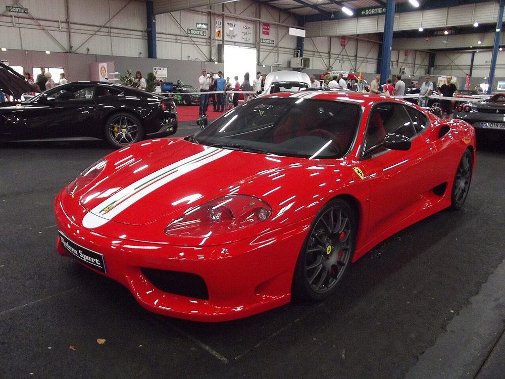 Red Ferrari 360 Challenge Stradale with white twin racing stripes parked indoors at a car show
