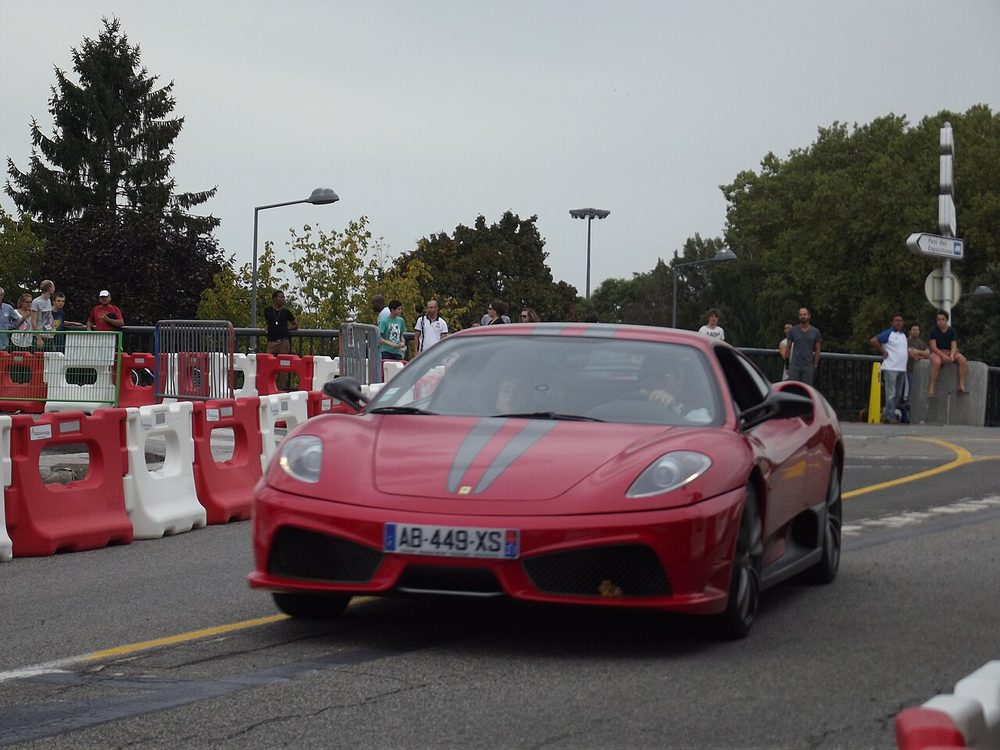 Red Ferrari 430 Scuderia with grey twin stripes on a road lined with barriers, seen from the front