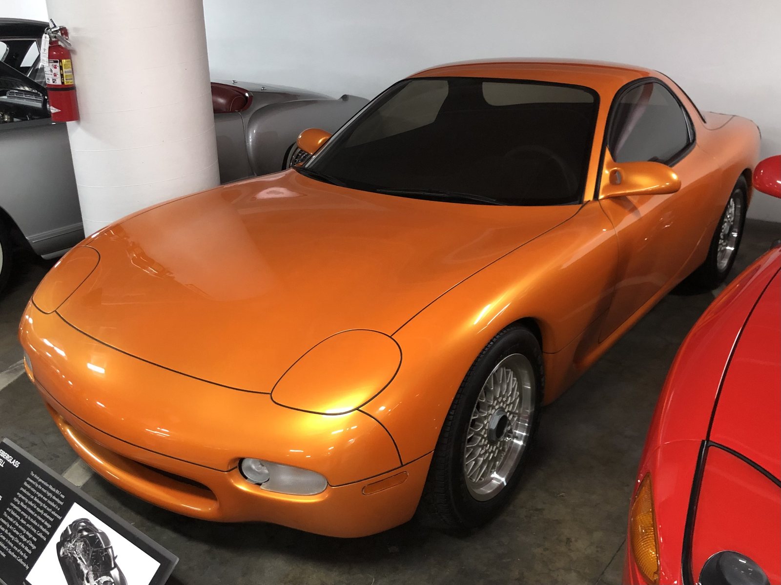 A glossy burnt-orange fiberglass concept model of the third-generation Mazda RX-7 parked among other cars on the concrete floor of the Petersen Museum's Vault, beside a white column and an information placard