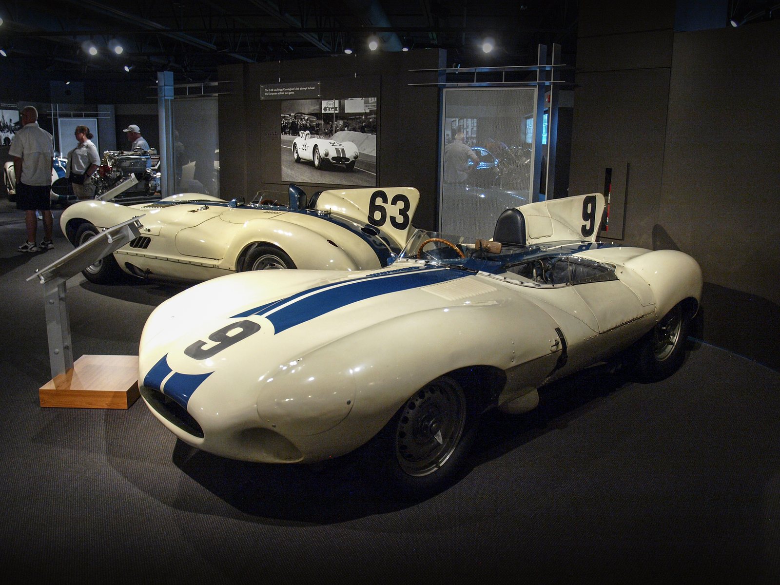 Two white racing cars with blue stripes and large race numbers 9 and 63 displayed without barriers in a softly lit gallery, the nearer one a 1955 Jaguar D-Type, with a black-and-white period racing photo on the wall behind