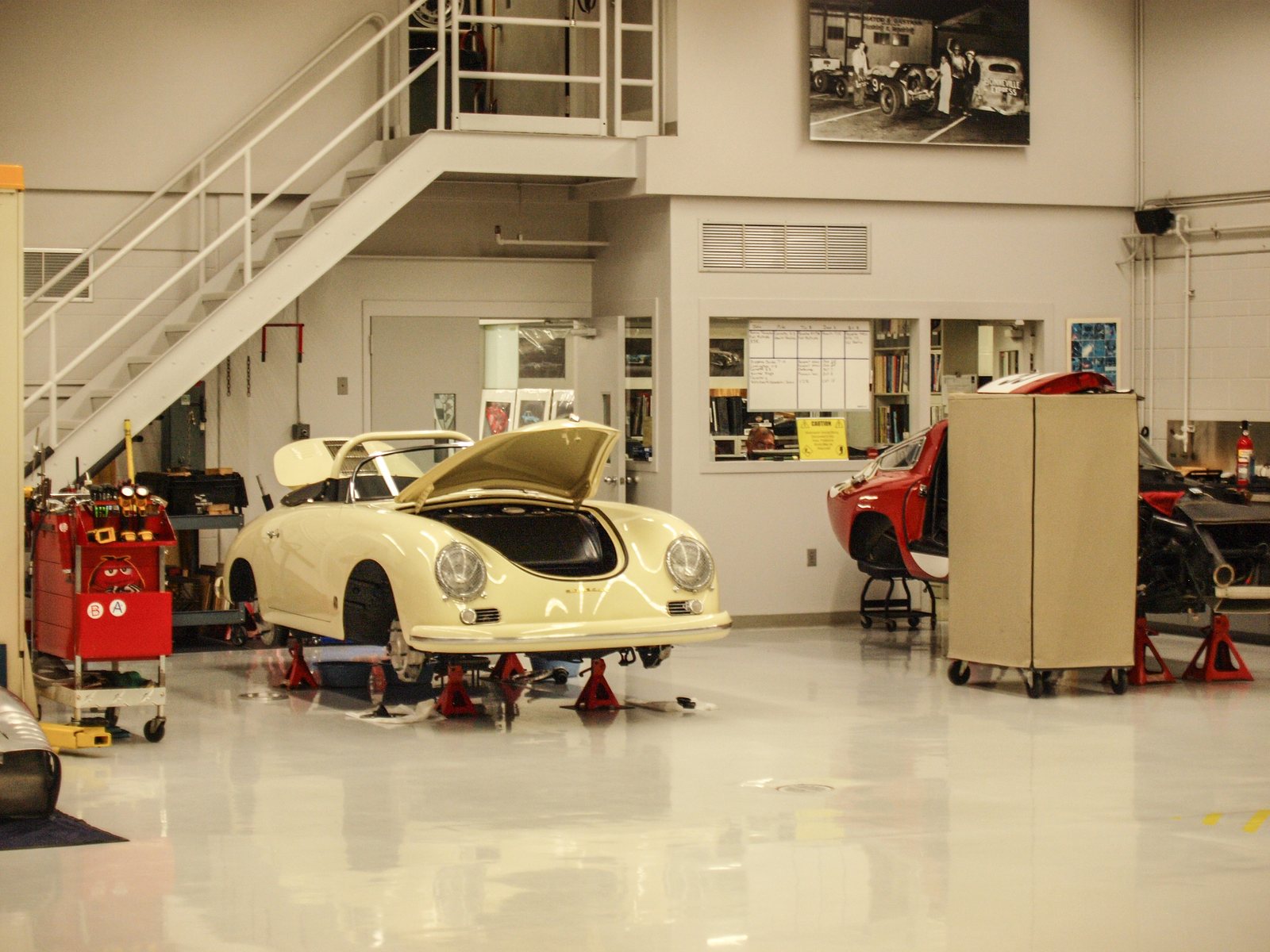 A bright, spotless restoration workshop with a white-tiled floor, where a cream Porsche 356 Speedster sits on red jack stands with its front lid open, a red tool cart beside it and a partially disassembled red car behind a partition