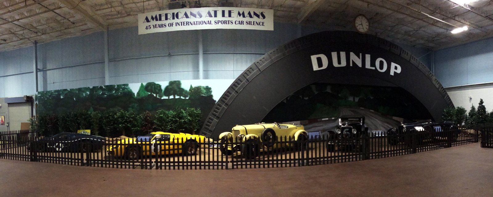 Panoramic view of the Americans at Le Mans exhibit at the Simeone Foundation Automotive Museum, with a row of racing cars including Ford GT40s and pre-war Stutz and duPont roadsters lined up in front of a painted forest backdrop and a replica of the famous Dunlop bridge
