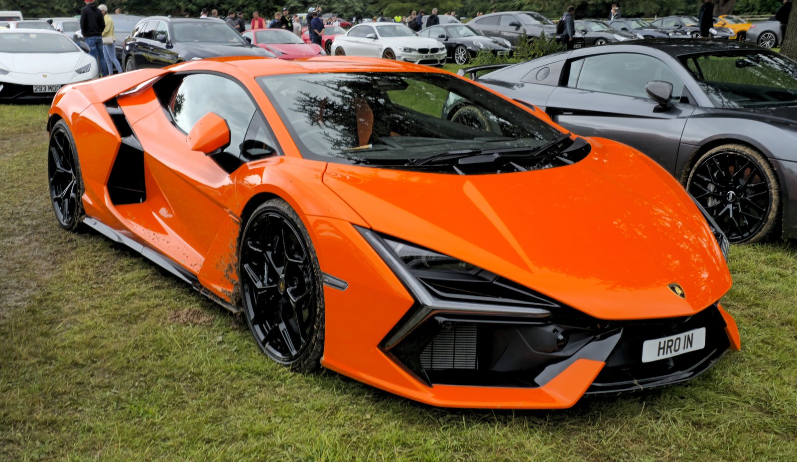Bright orange Lamborghini Revuelto seen from the front three-quarter angle on grass at a car show, showing its sharp nose, Y-shaped headlights, black wheels and large side intakes