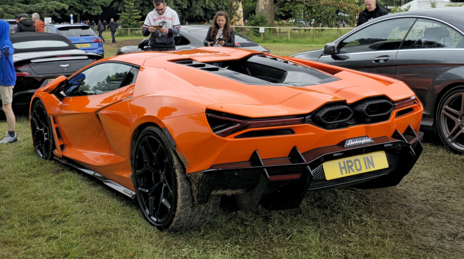Rear three-quarter view of an orange Lamborghini Revuelto at an outdoor car show, showing the exposed V12 engine bay behind the cabin, the hexagonal twin exhaust outlets and the rear diffuser